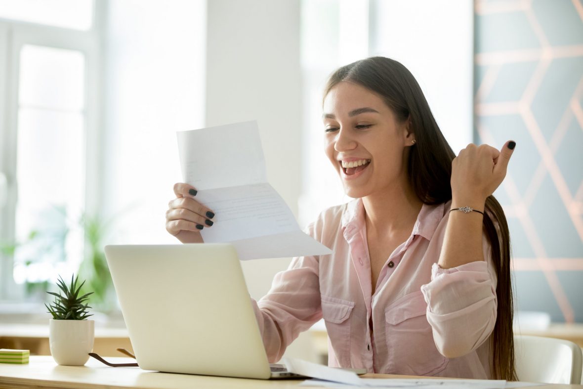 Jente smiler og leser papirbrev foran laptop