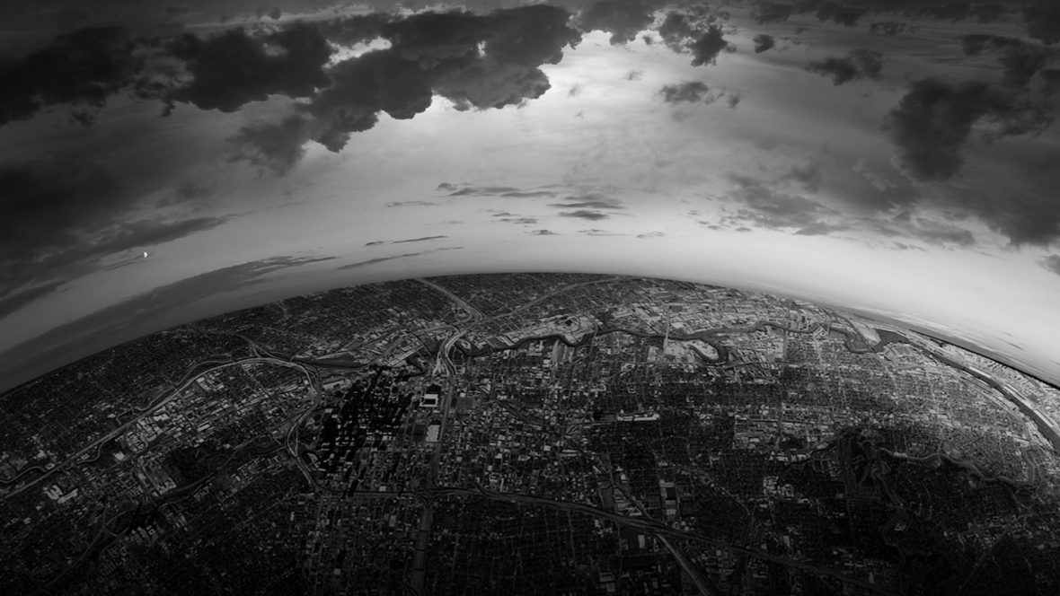 Sort/hvitt-bilde tatt høyt ovenfra, hvor jorden og en by kurver rundt på sidene og skyene er mørke på himmelen