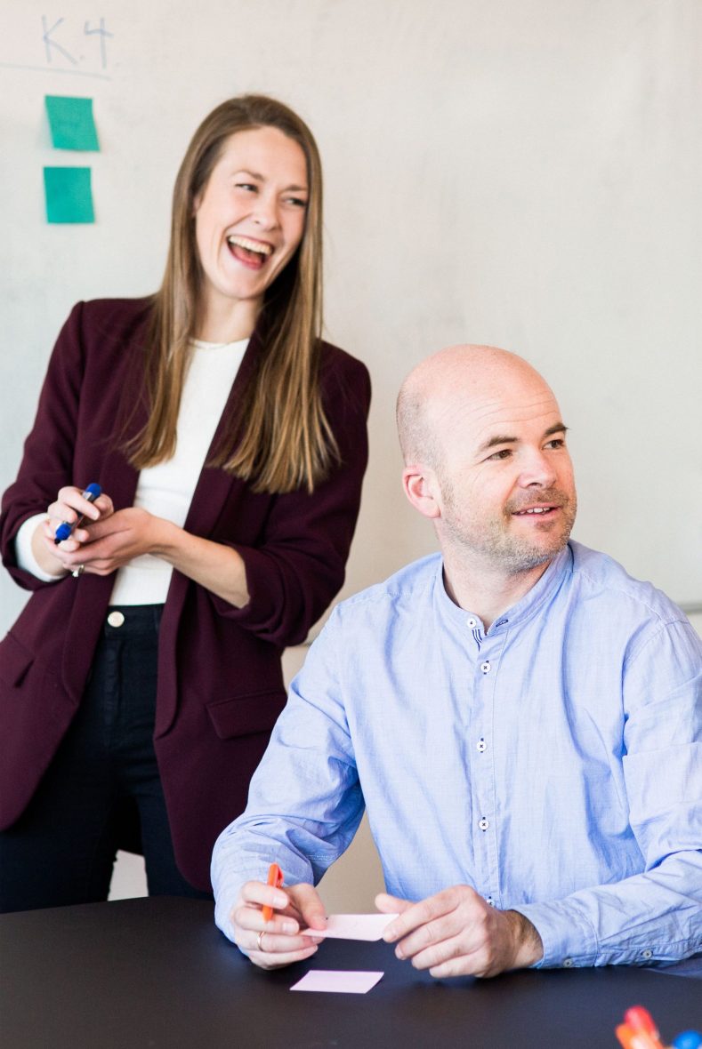 Kvinne står og ler ved siden av mann som skriver på post-it lapp