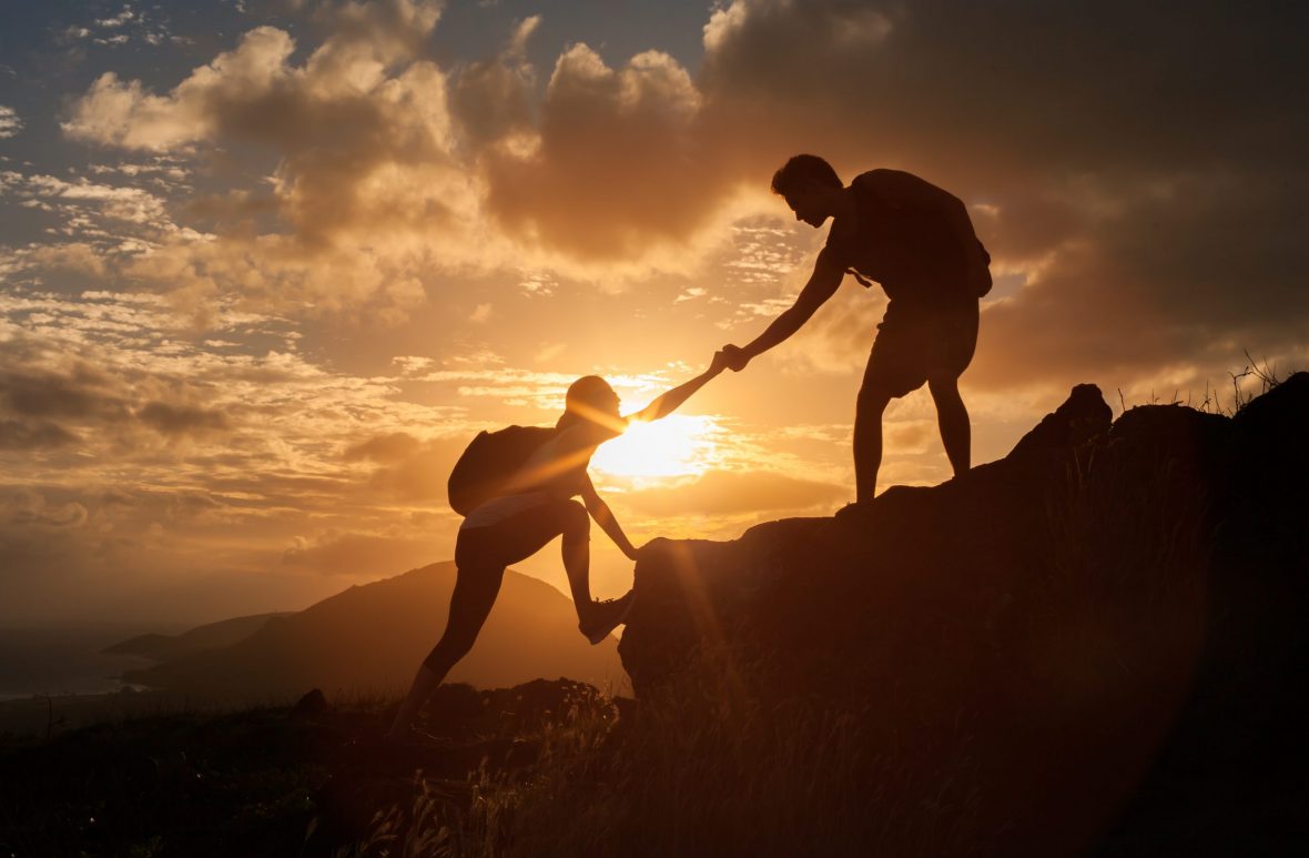 Siluett av to mennesker som hjelper hverandre opp et fjell i solnedgang.